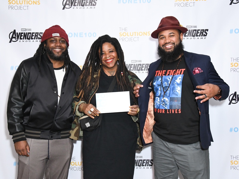 Three people in front of a step-and-repeat sign with The Solutions Project and Avengers logos on it
